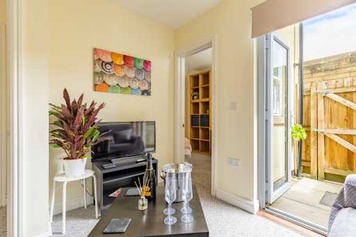 Bright living room with a television, seating, coffee table, shelving, plant and open doors to a small fenced patio.