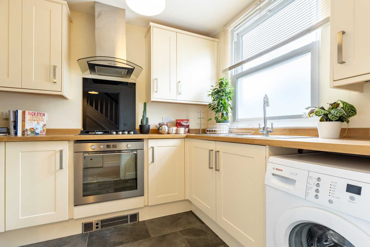 Bright fitted kitchen with cream cabinets, an oven, sink, large window, washing machine and tiled floor.