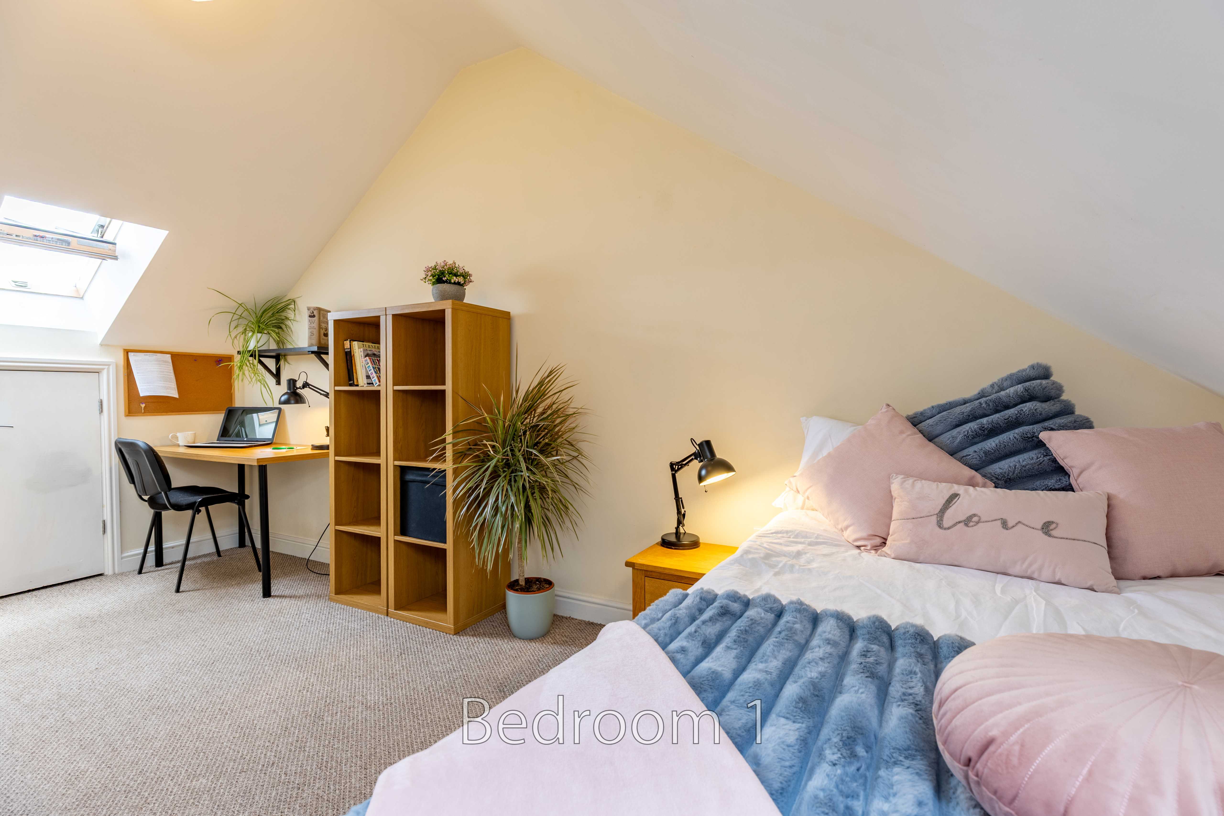 A furnished attic bedroom with a bed, desk, chair, shelving and skylight.