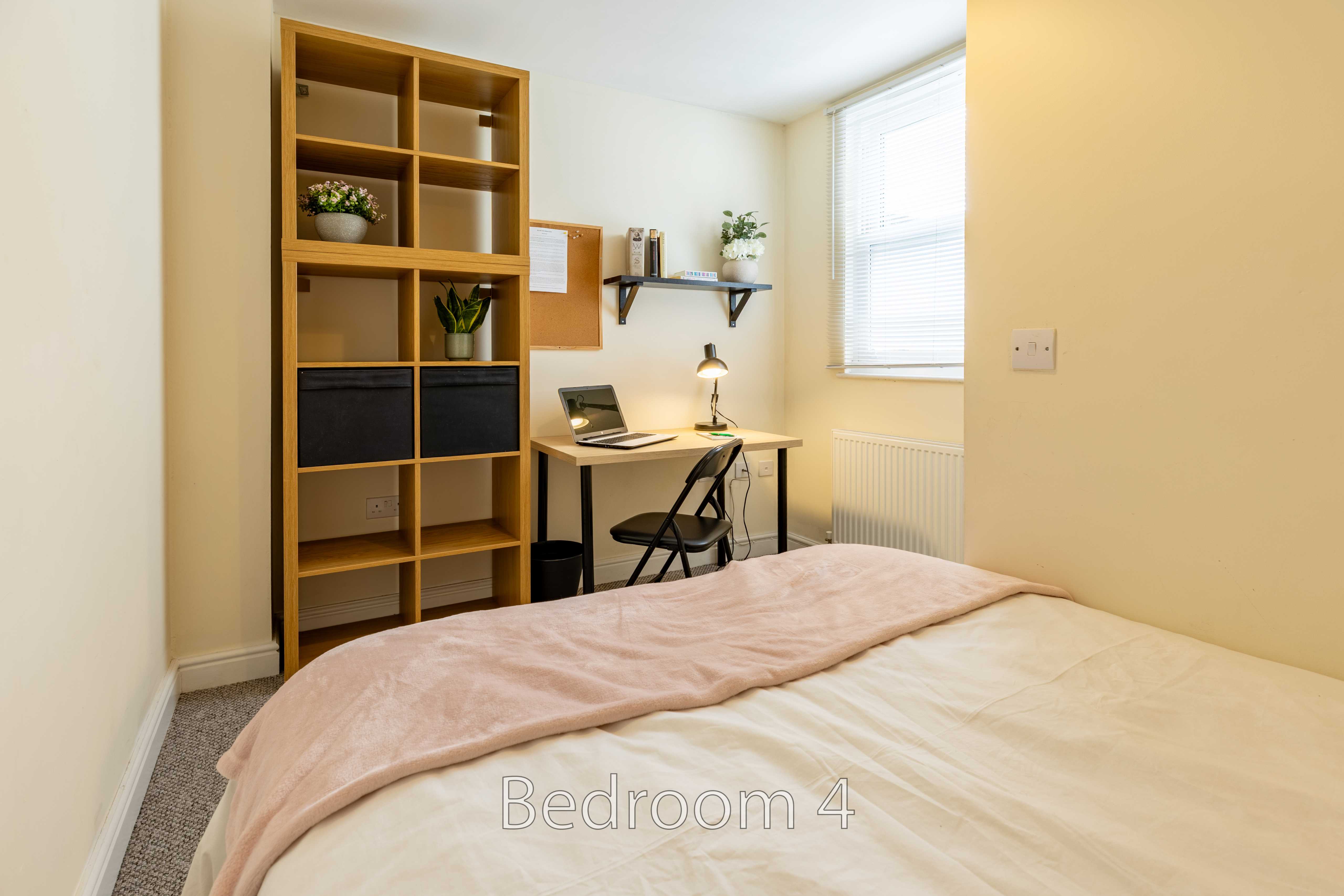 A bedroom with a bed, desk, chair, shelving unit, window and radiator.
