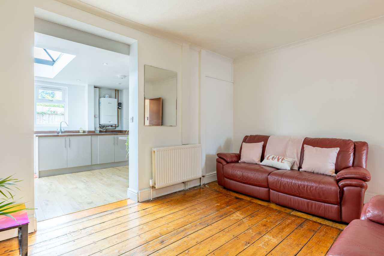 Living room with a red leather sofa, wooden floorboards, radiator and an open doorway to a bright kitchen.