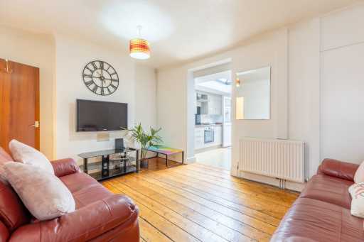 Bright living room with wood flooring, two leather sofas, a wall-mounted television, a clock, a radiator and an open doorway to the kitchen.