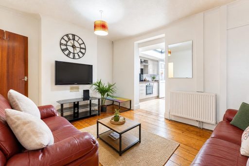 Bright living room with wood flooring, two leather sofas, a wall-mounted television, a clock, a radiator and an open doorway to the kitchen.
