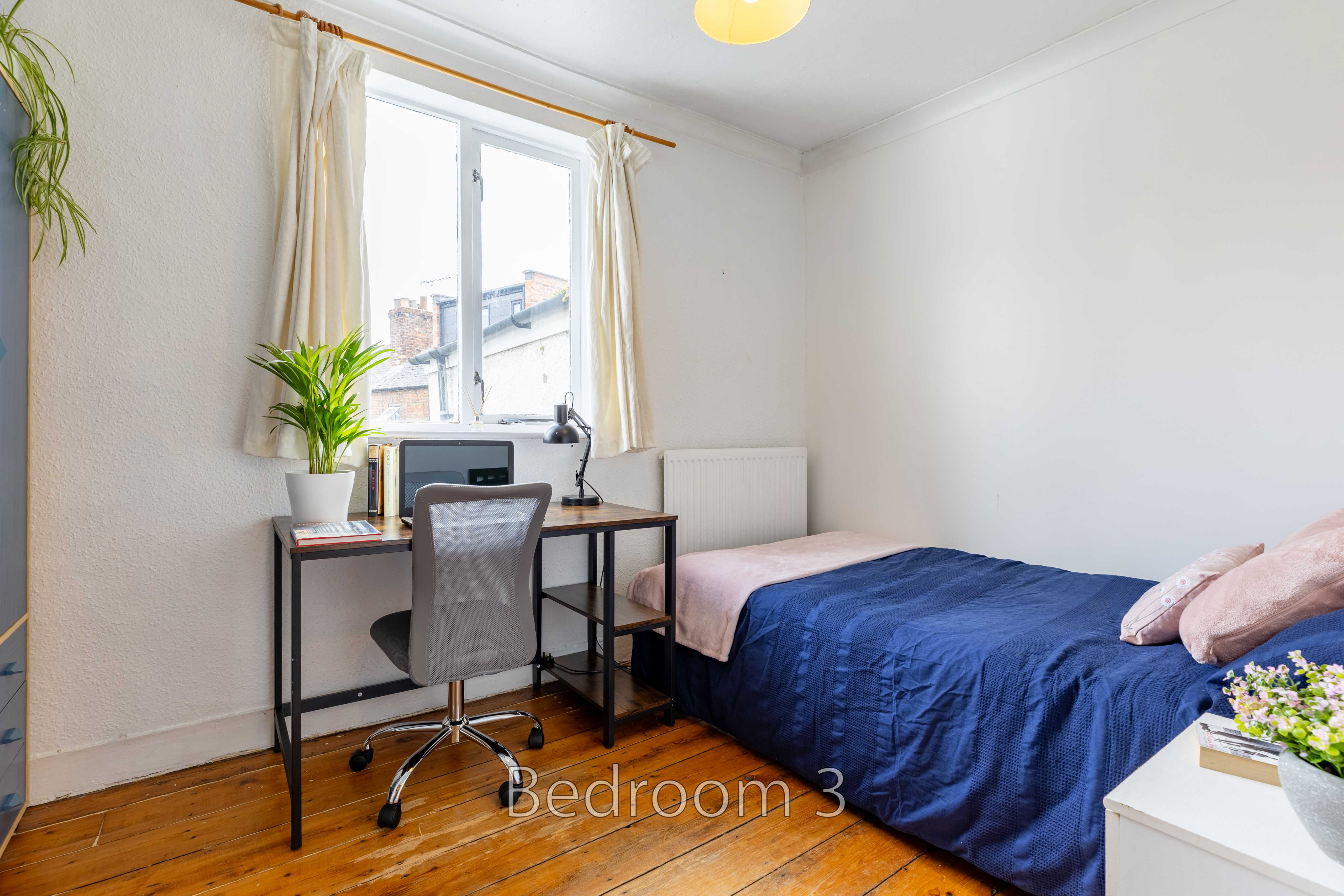 A bedroom with a bed, desk, chair, window, curtains and wooden flooring.