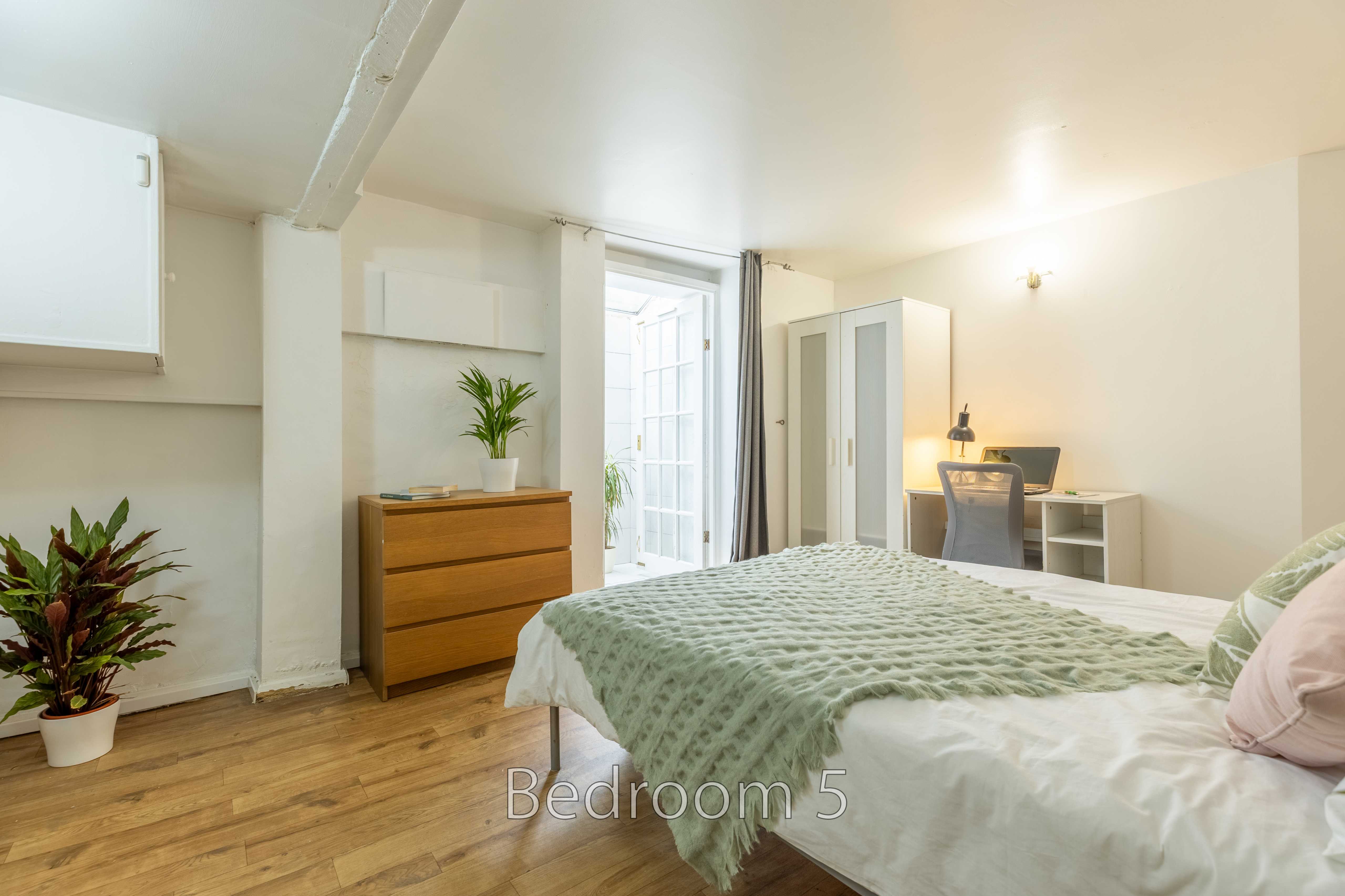 A furnished bedroom with a double bed, chest of drawers, wardrobe, desk and potted plants.
