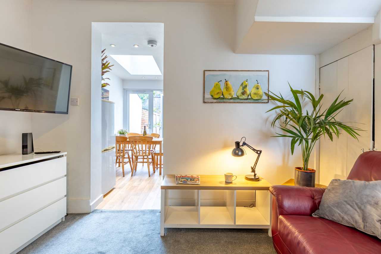 Bright living area with a red sofa, media unit, coffee table, plant and dining area visible through an open doorway.