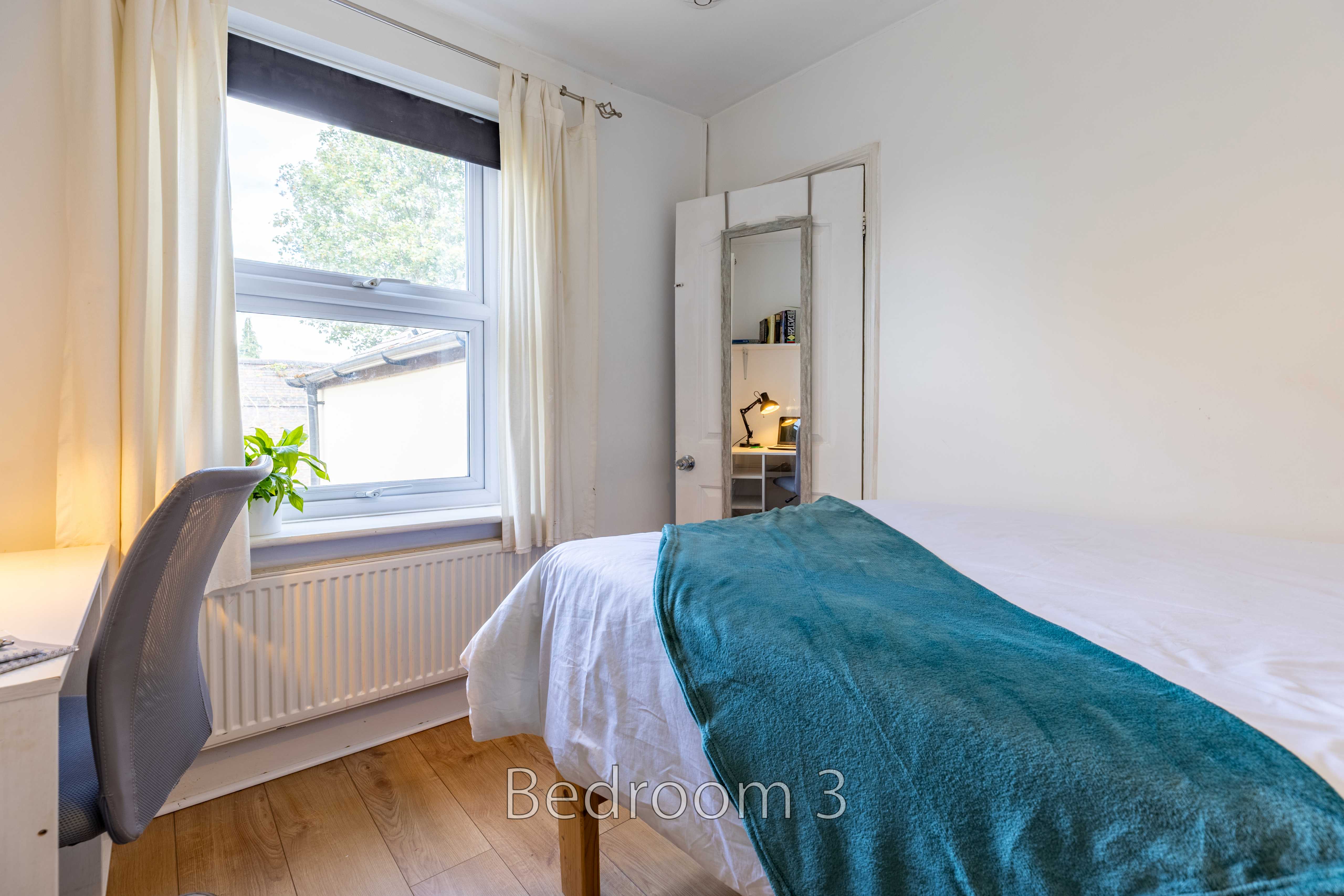 A bedroom with a bed, window, curtains, radiator, desk chair and wooden floor.