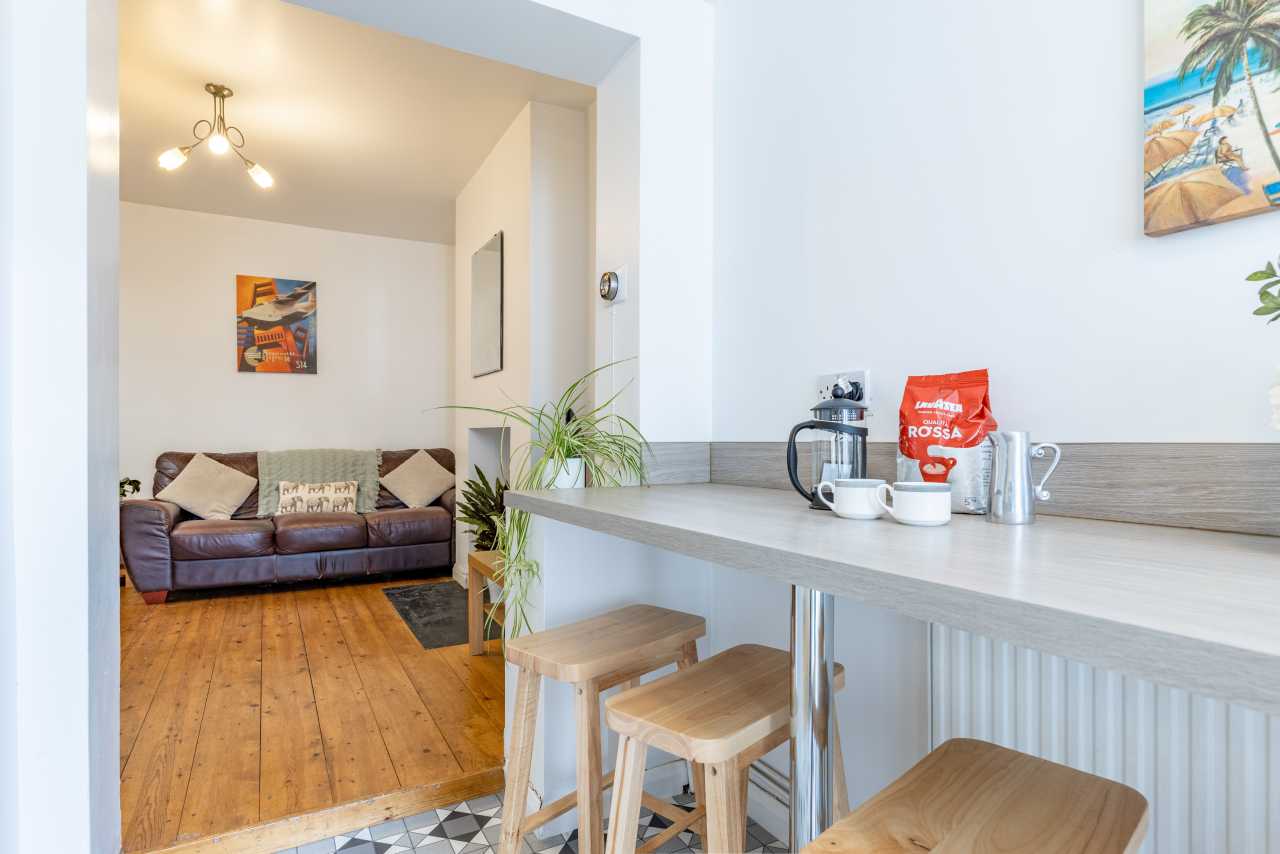 Open-plan living area with a sofa, wood flooring, kitchen counter, stools and coffee-making items.