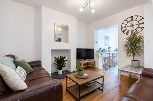 Bright living room with leather sofas, a television, wooden floor, fireplace, plants and a dining area visible through a doorway.