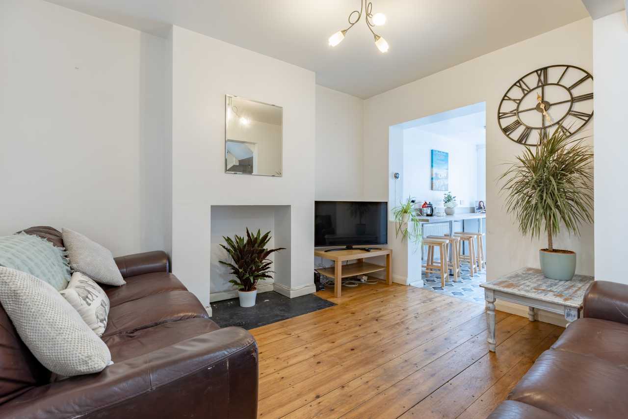 Bright living room with leather sofas, a television, wooden floor, fireplace, plants and a dining area visible through a doorway.