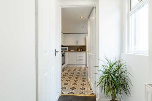 View through an open doorway into a bright kitchen with white cabinets, patterned floor tiles, a window, and a potted plant.