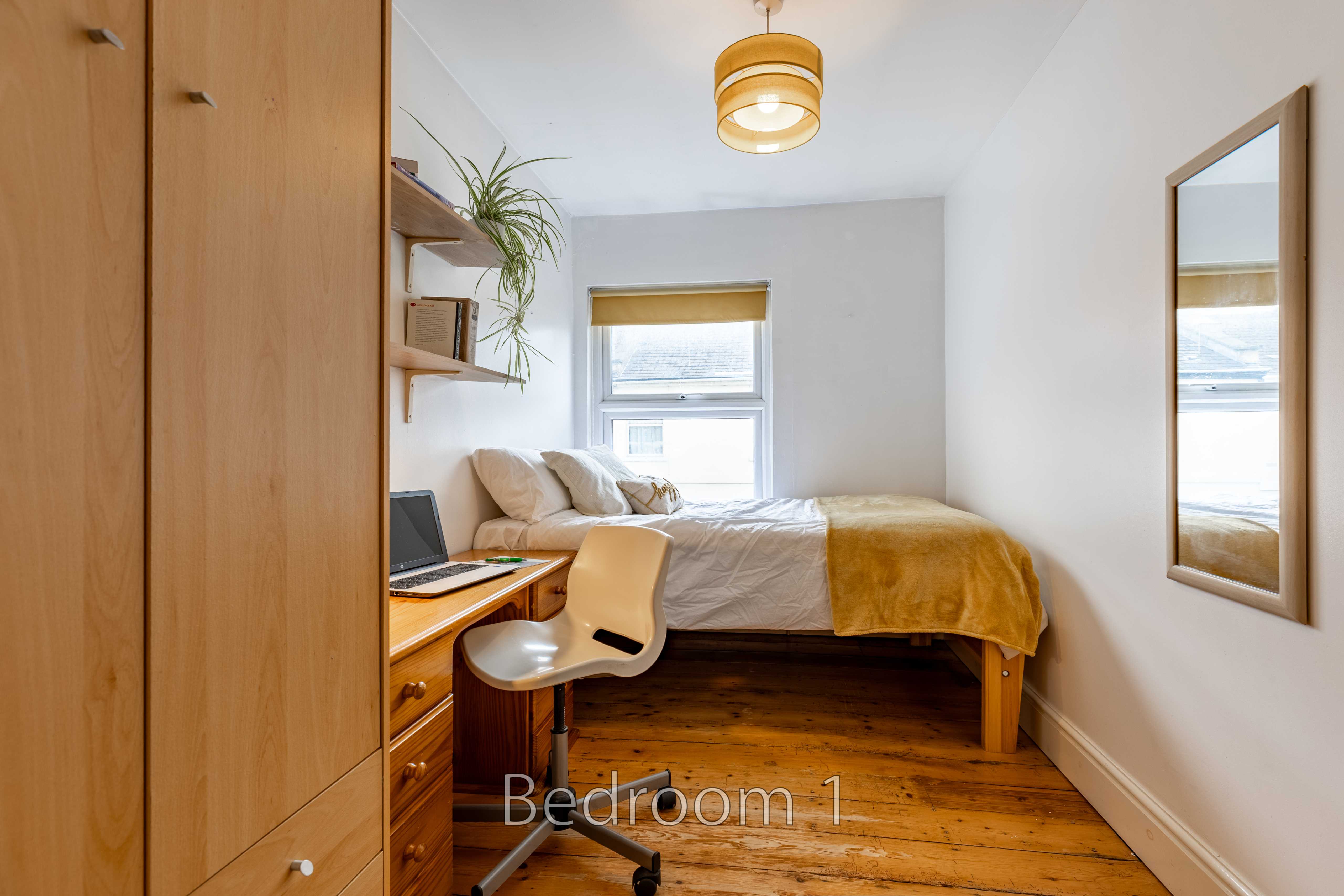 A furnished bedroom with a bed, desk and chair, wooden flooring, wardrobe, shelves and a window.