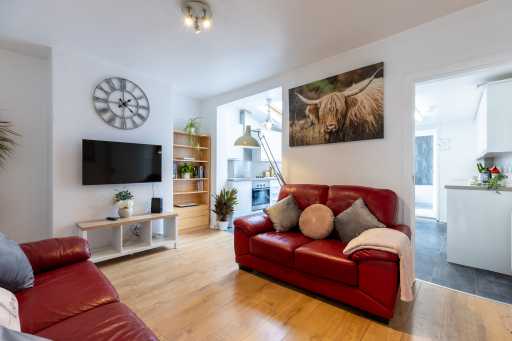 Bright living room with red leather sofas, a television, wooden flooring, shelving, wall art and an open kitchen area.