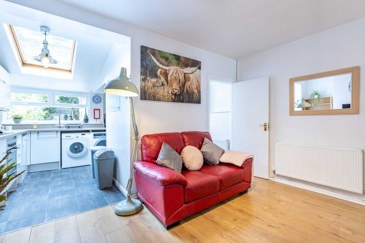 Open-plan living area with a red sofa, wooden flooring, radiator, wall art and an adjoining kitchen with skylight and appliances.