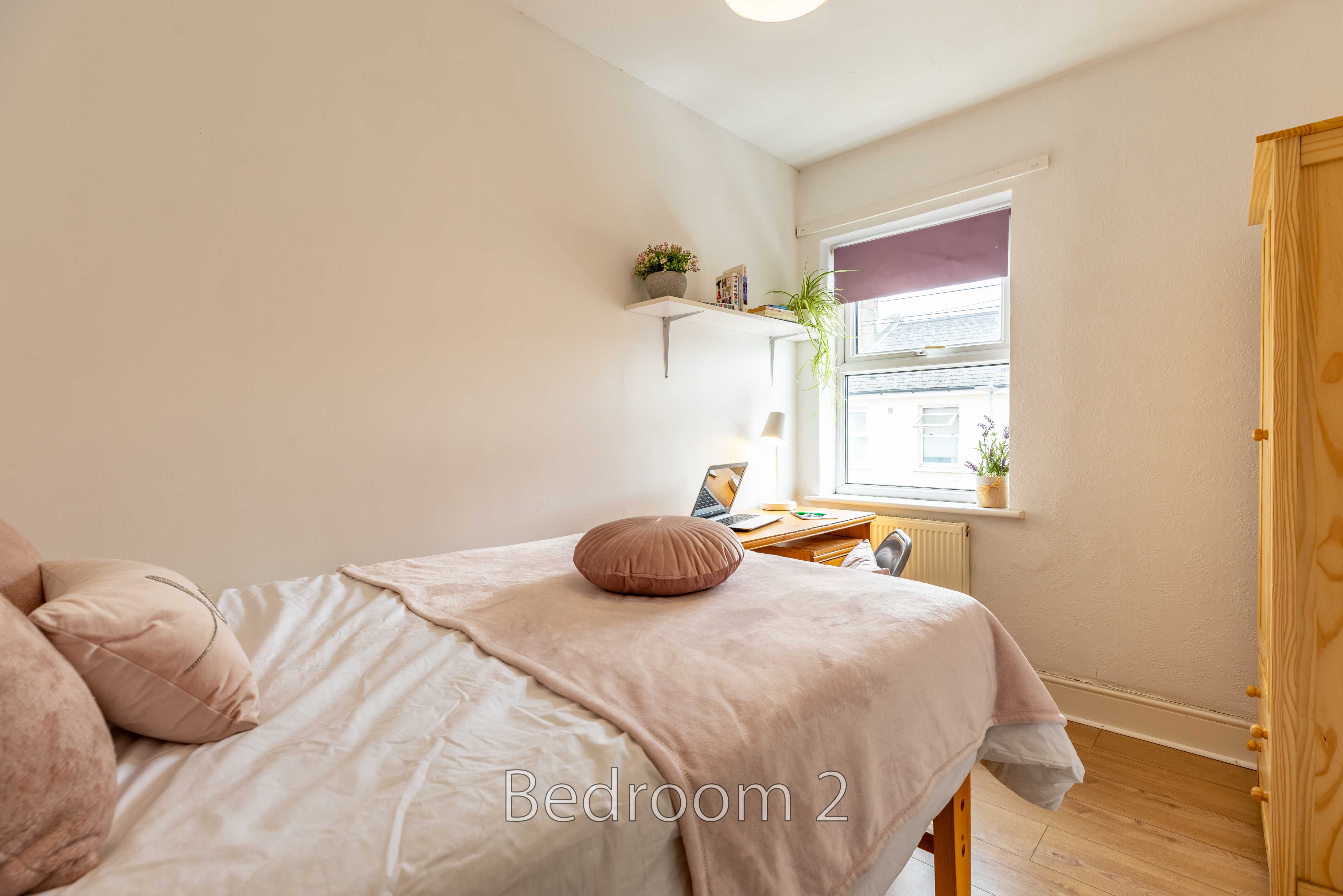 A bedroom with a double bed, pale bedding, a window, plants on a shelf, and a wooden wardrobe.