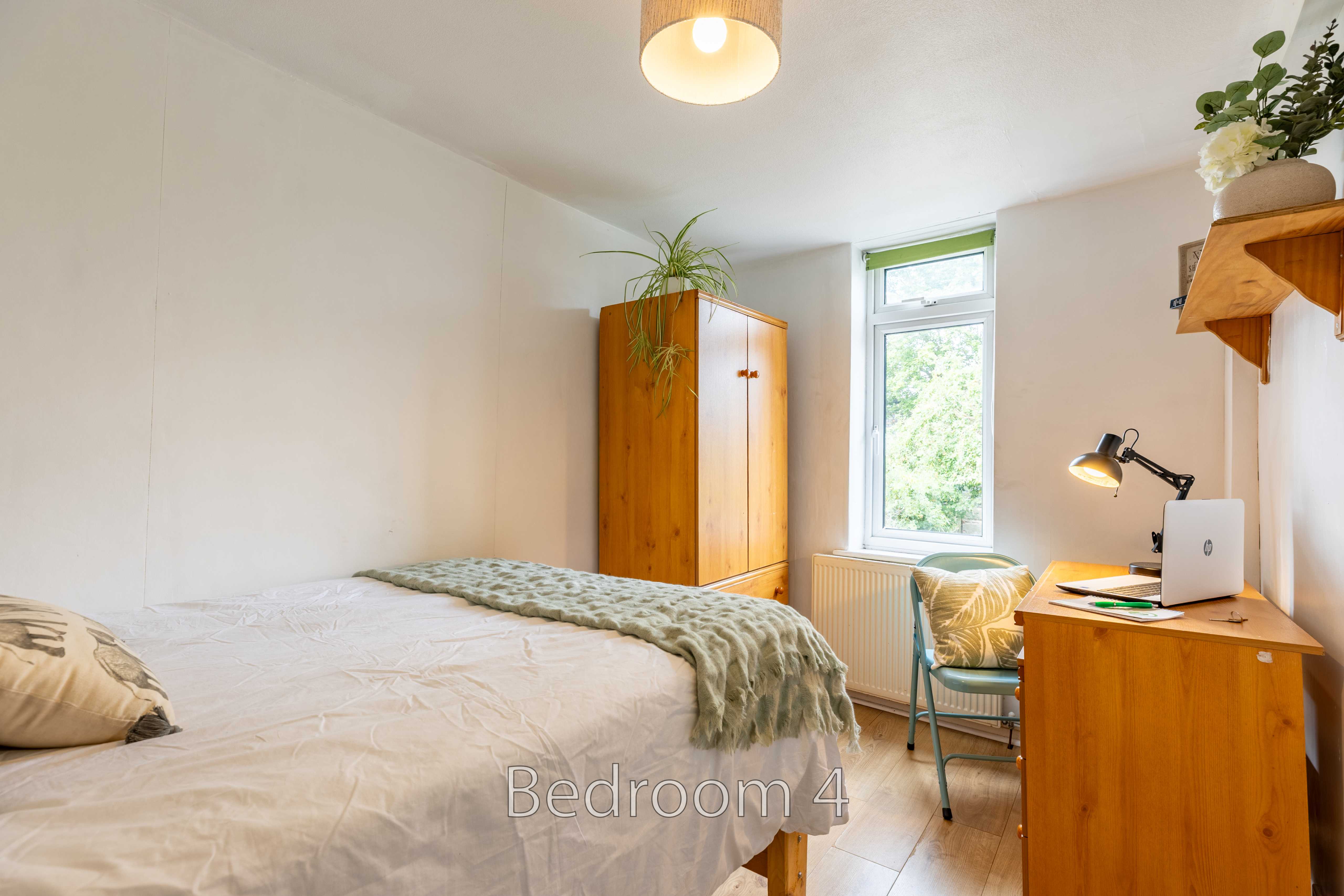 A furnished bedroom with a bed, wooden wardrobe, desk, chair, window and plants.