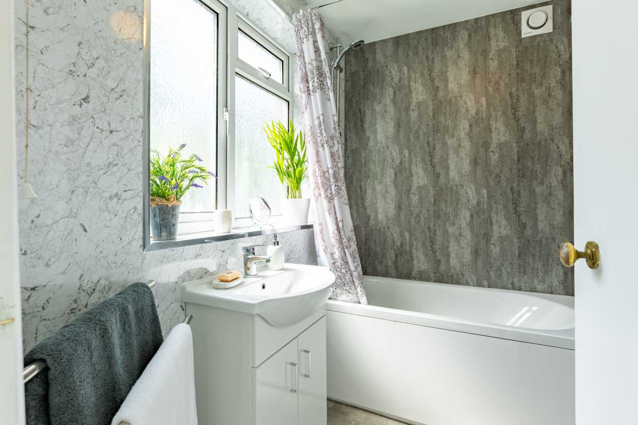 A bright bathroom with a bathtub, washbasin, window, towel rail and patterned wall coverings.