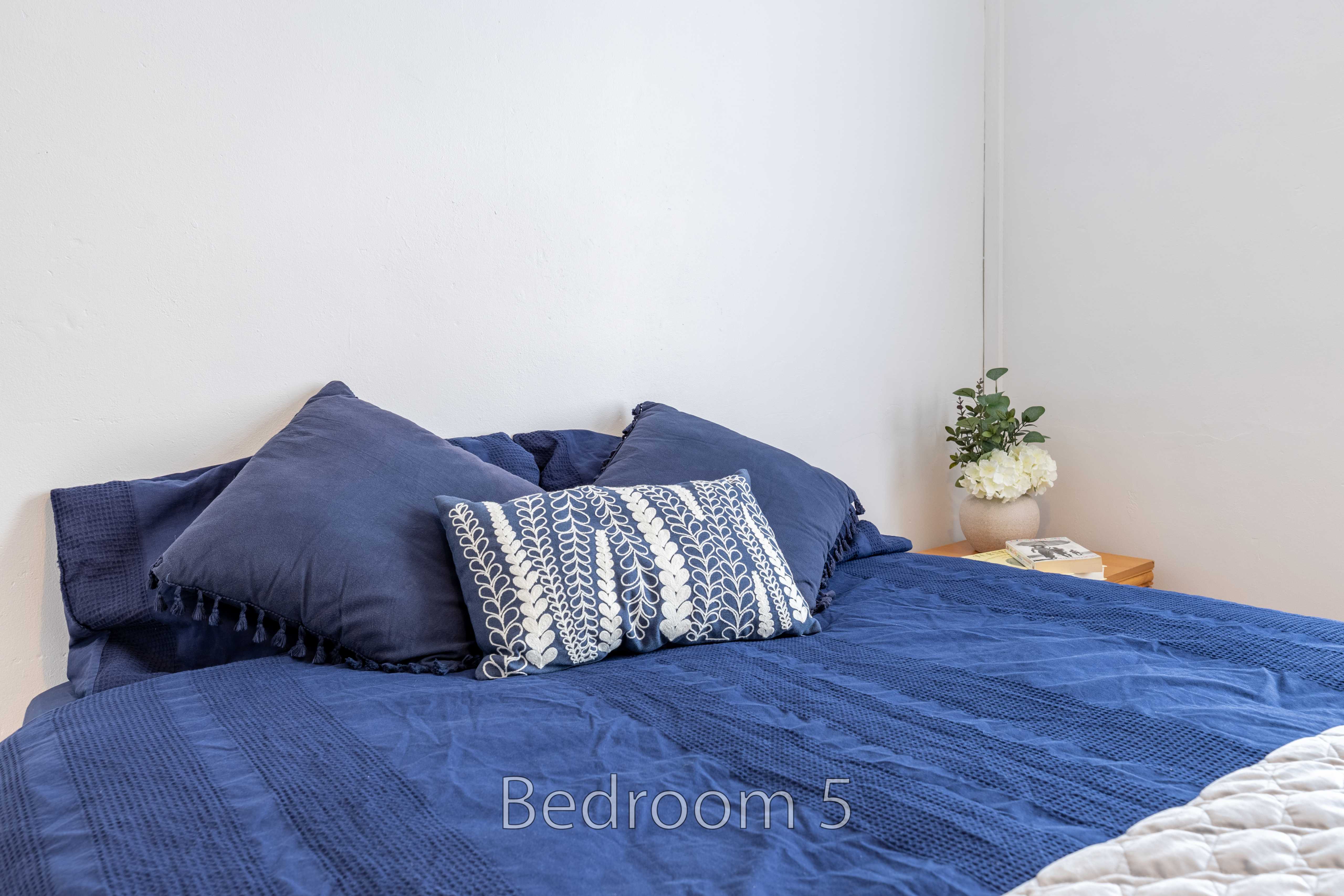 A bedroom with a blue bedspread, blue pillows, a bedside table and a vase of flowers.