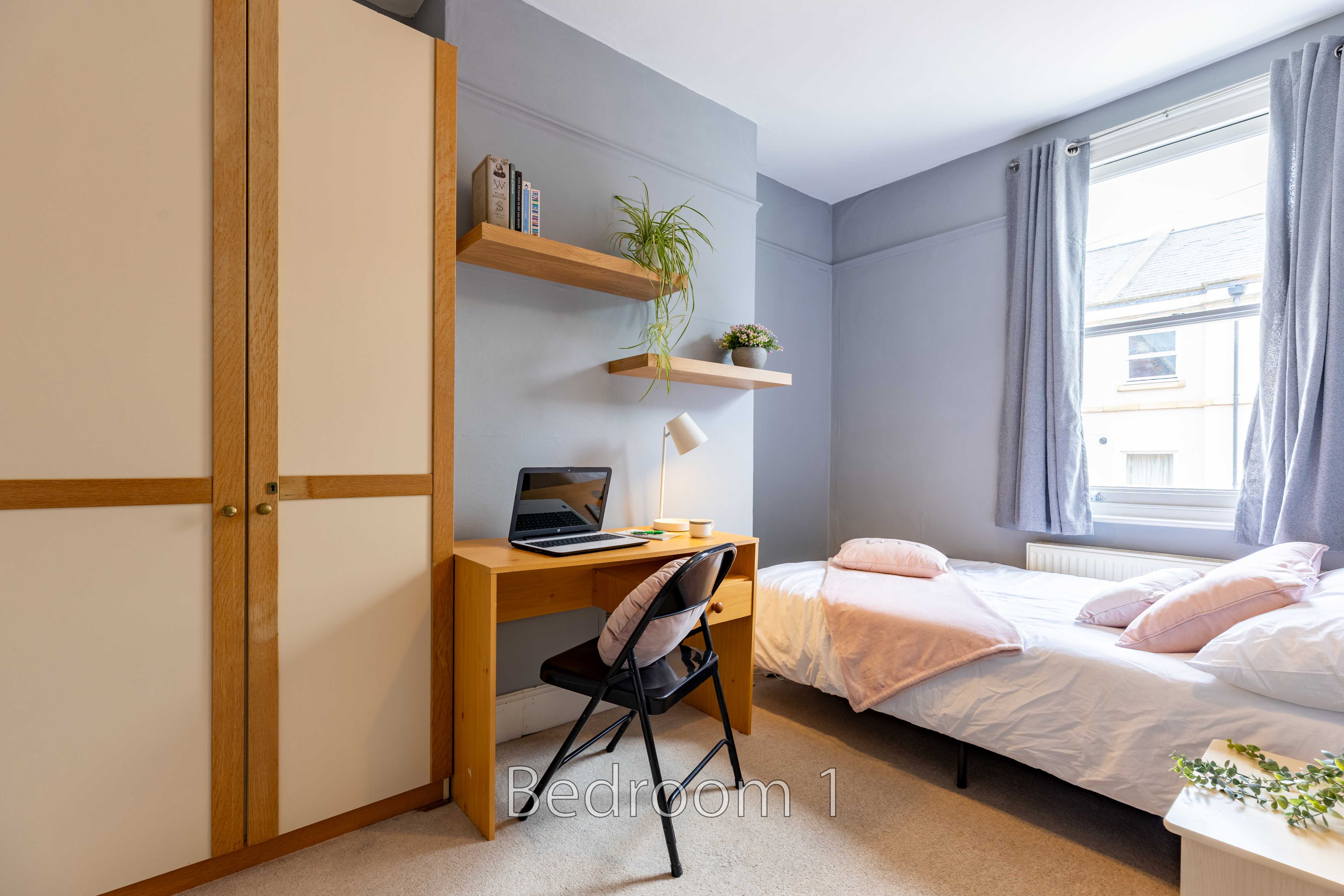 A furnished bedroom with two single beds, a desk, wardrobe, shelves and a window.