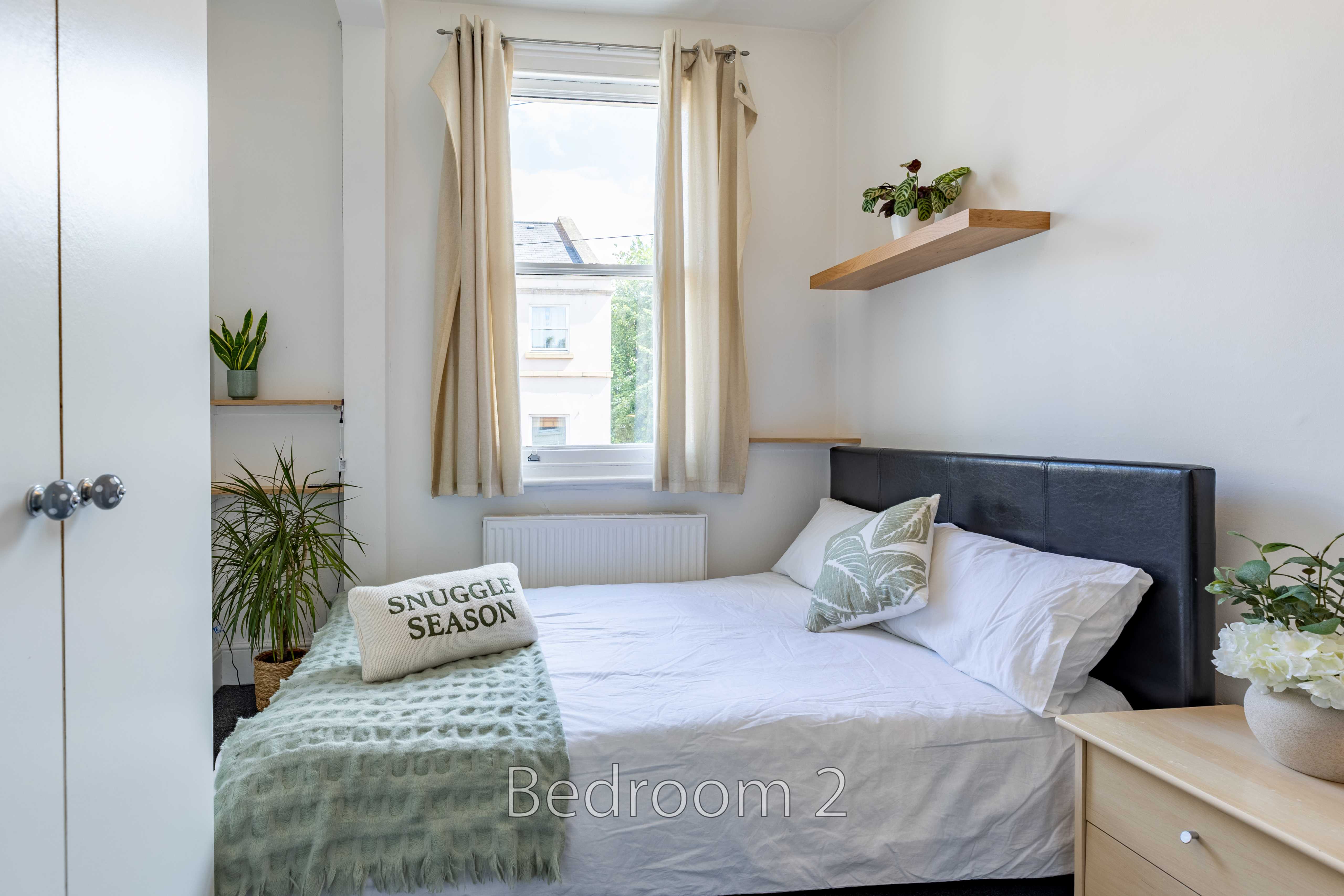 A furnished bedroom with a double bed, window, curtains, plants and wall shelves.
