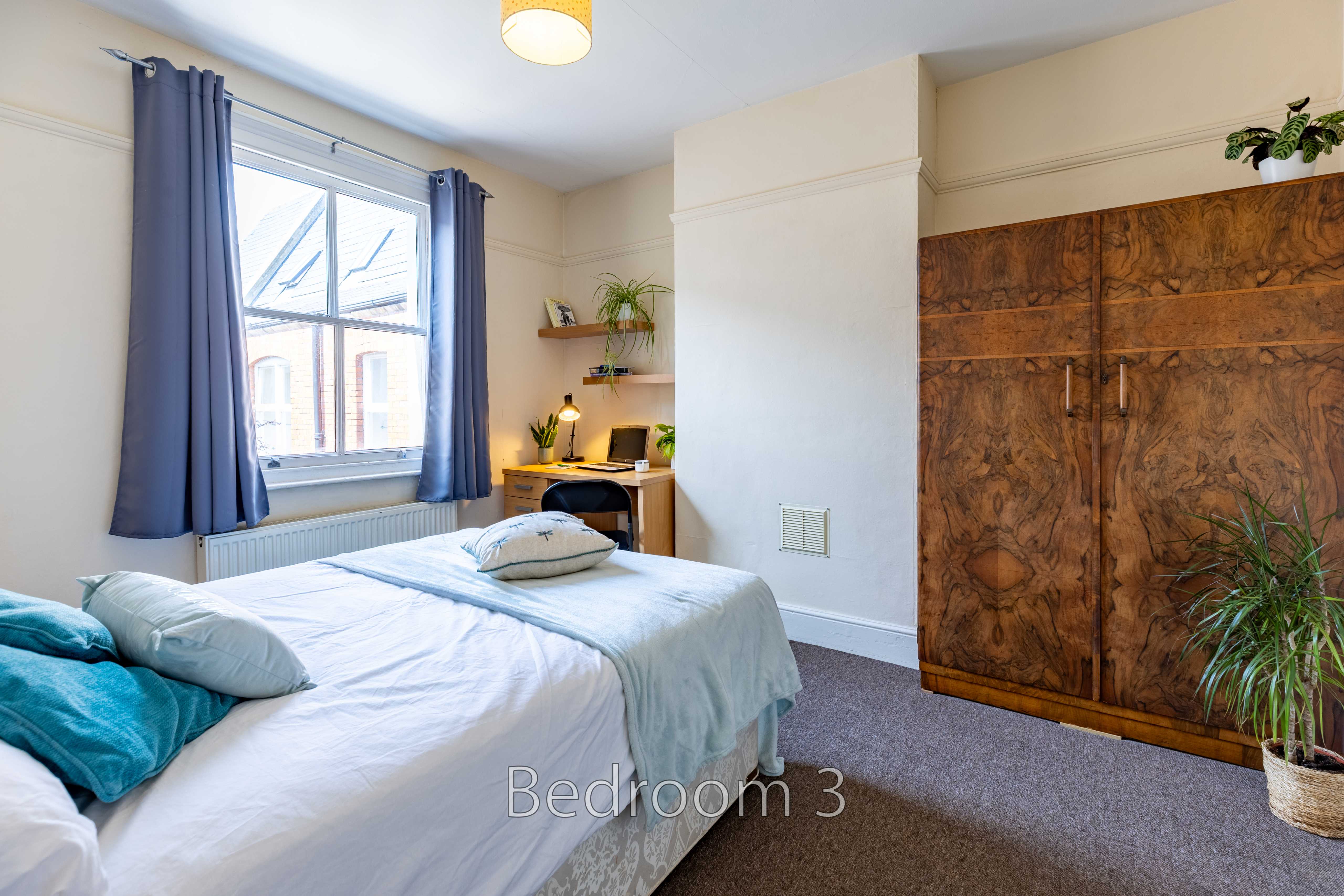 A furnished bedroom with a bed, window, desk, curtains and a large wooden wardrobe.