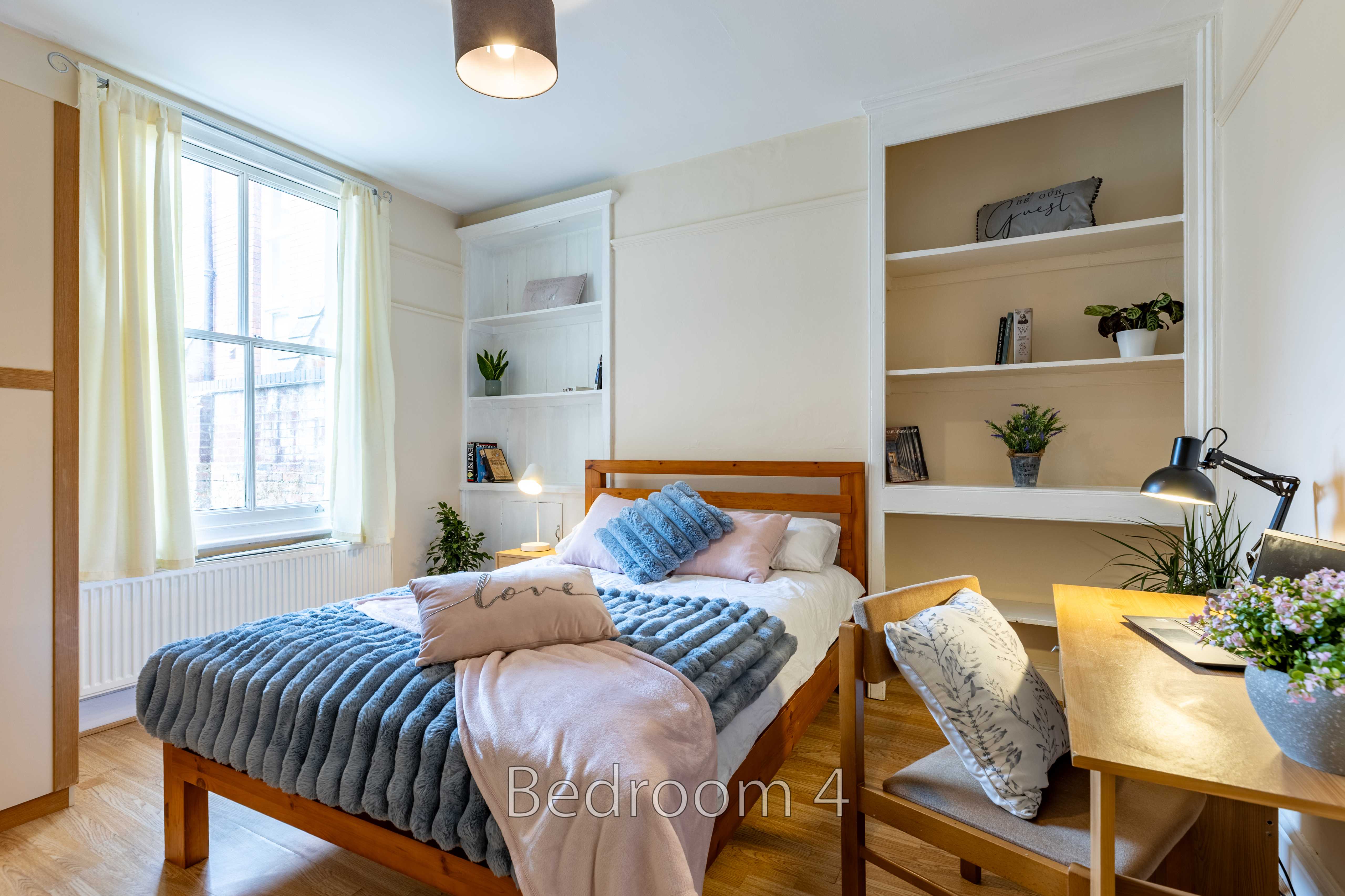 A bright bedroom with a double bed, desk, chair, built-in shelves, and a large window.