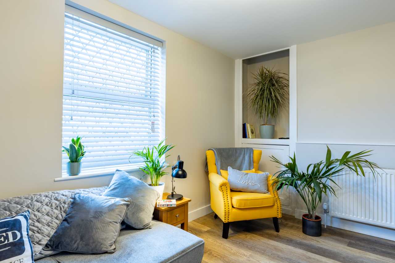 Bright residential room with a grey sofa, yellow armchair, plants, window blinds and a radiator.