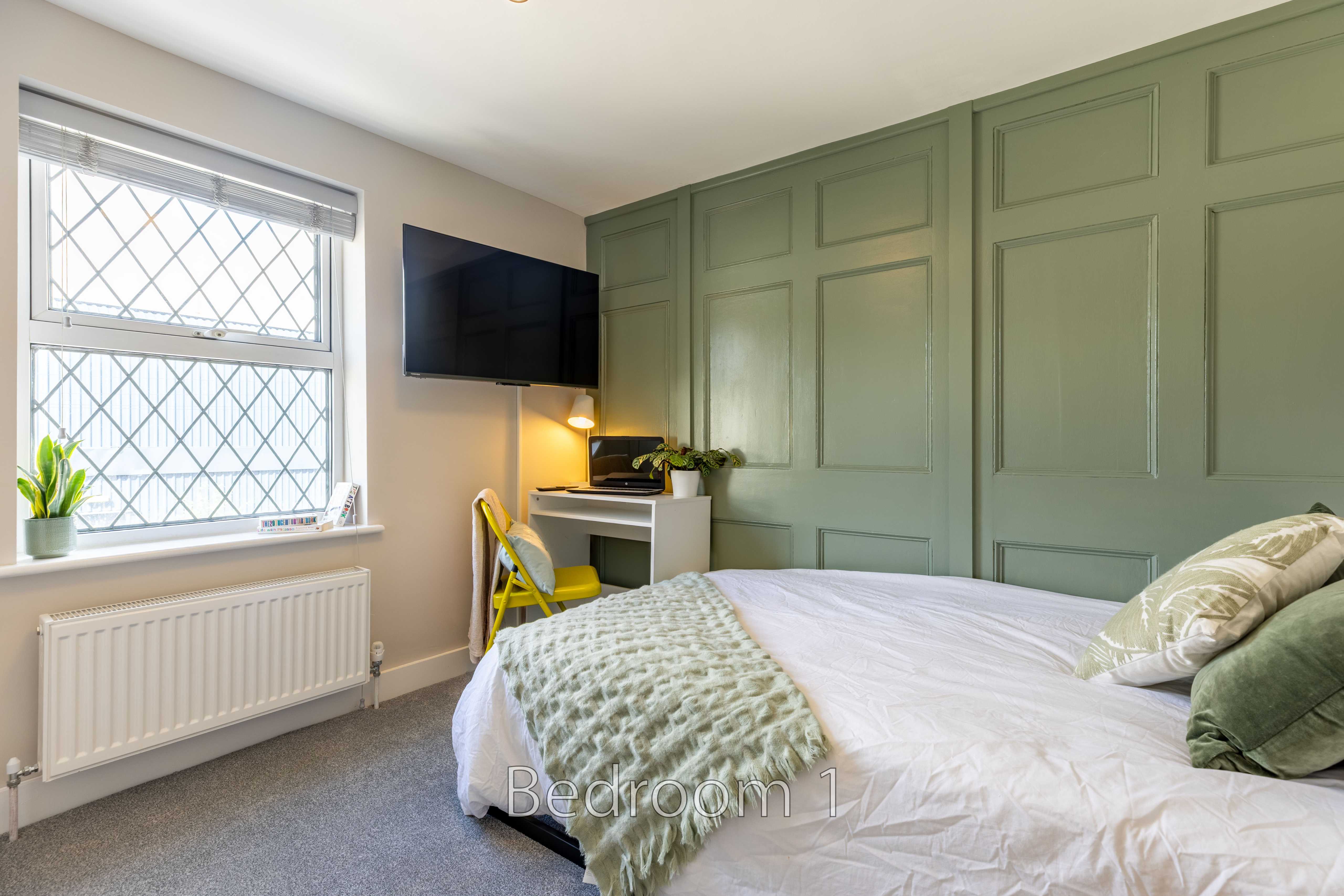A bedroom with a bed, green paneled wall, television, desk, window and radiator.
