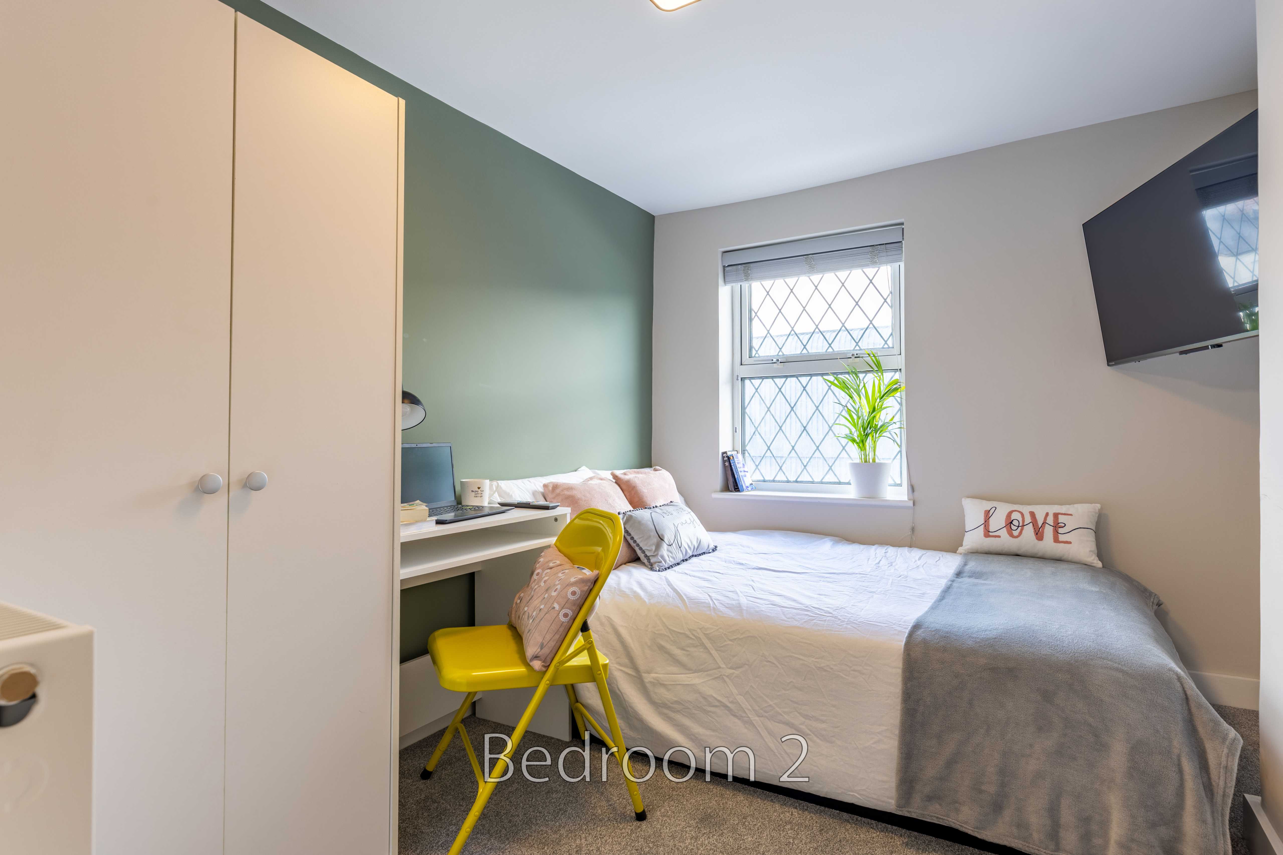 A single bedroom with a bed, desk, chair, wardrobe, window and wall-mounted television.