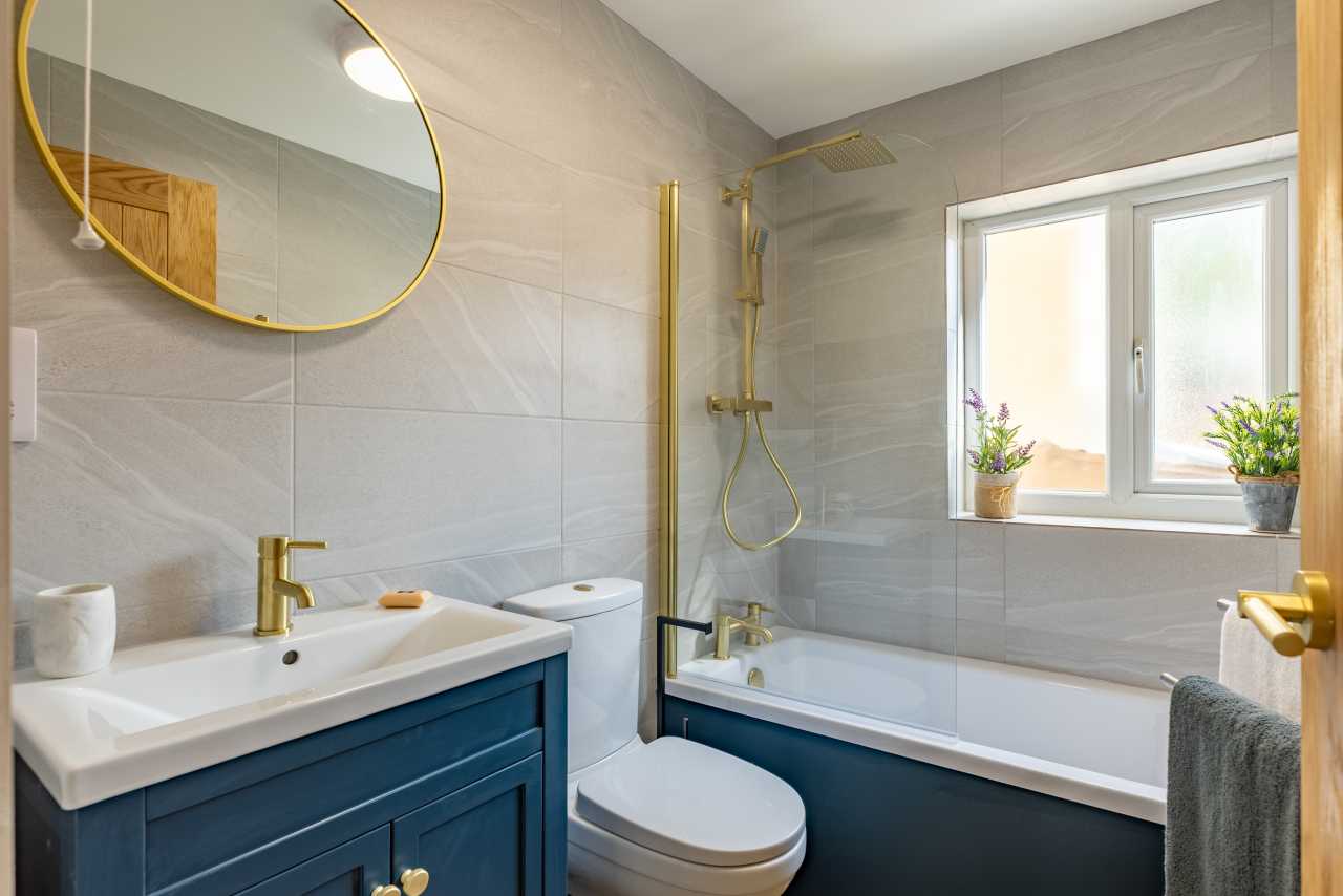 Bright modern bathroom with a bathtub, glass shower screen, toilet, sink with blue vanity, round mirror, and frosted window.