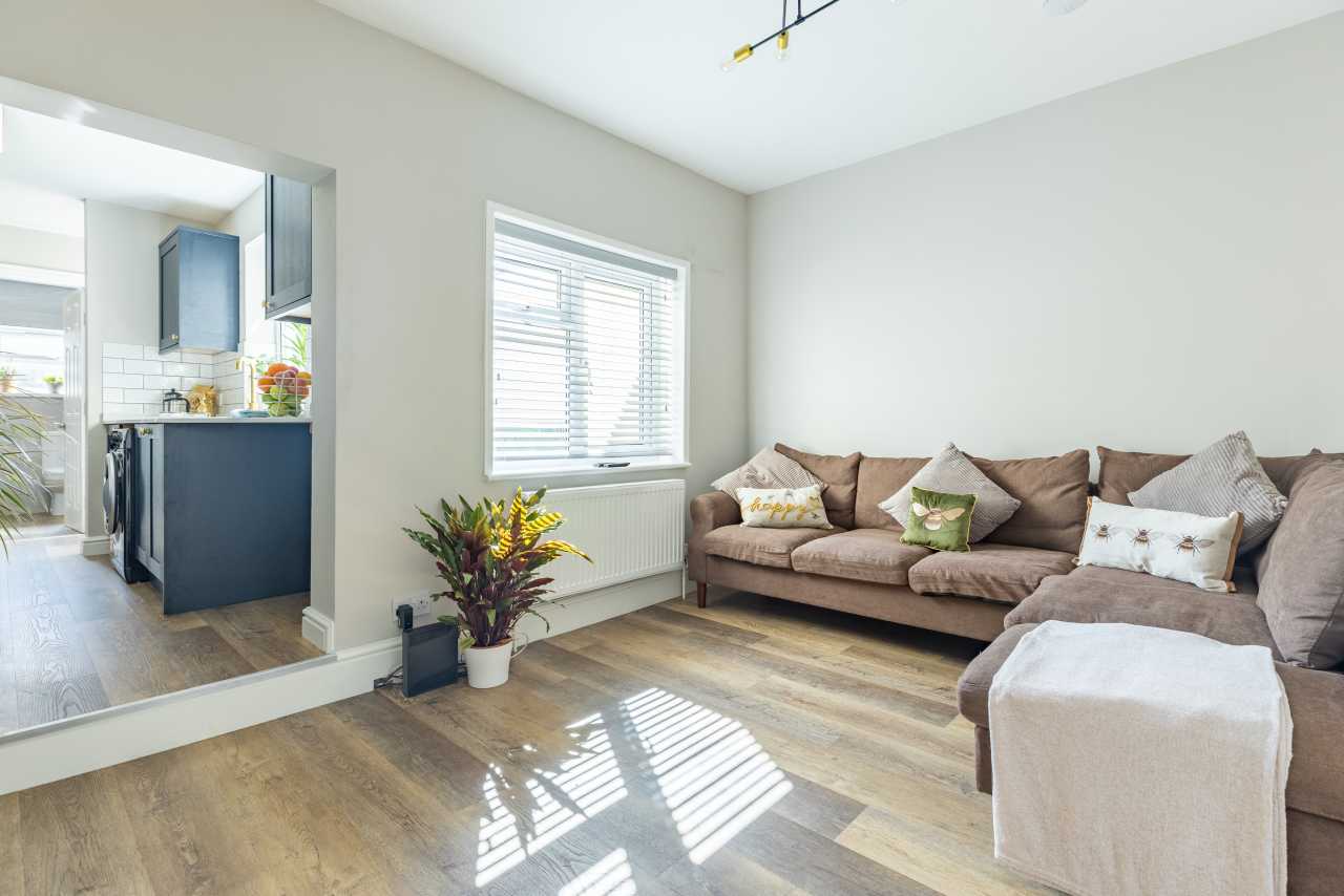 Bright living room with sofas, a window with blinds, wooden flooring, and an adjoining kitchen area.