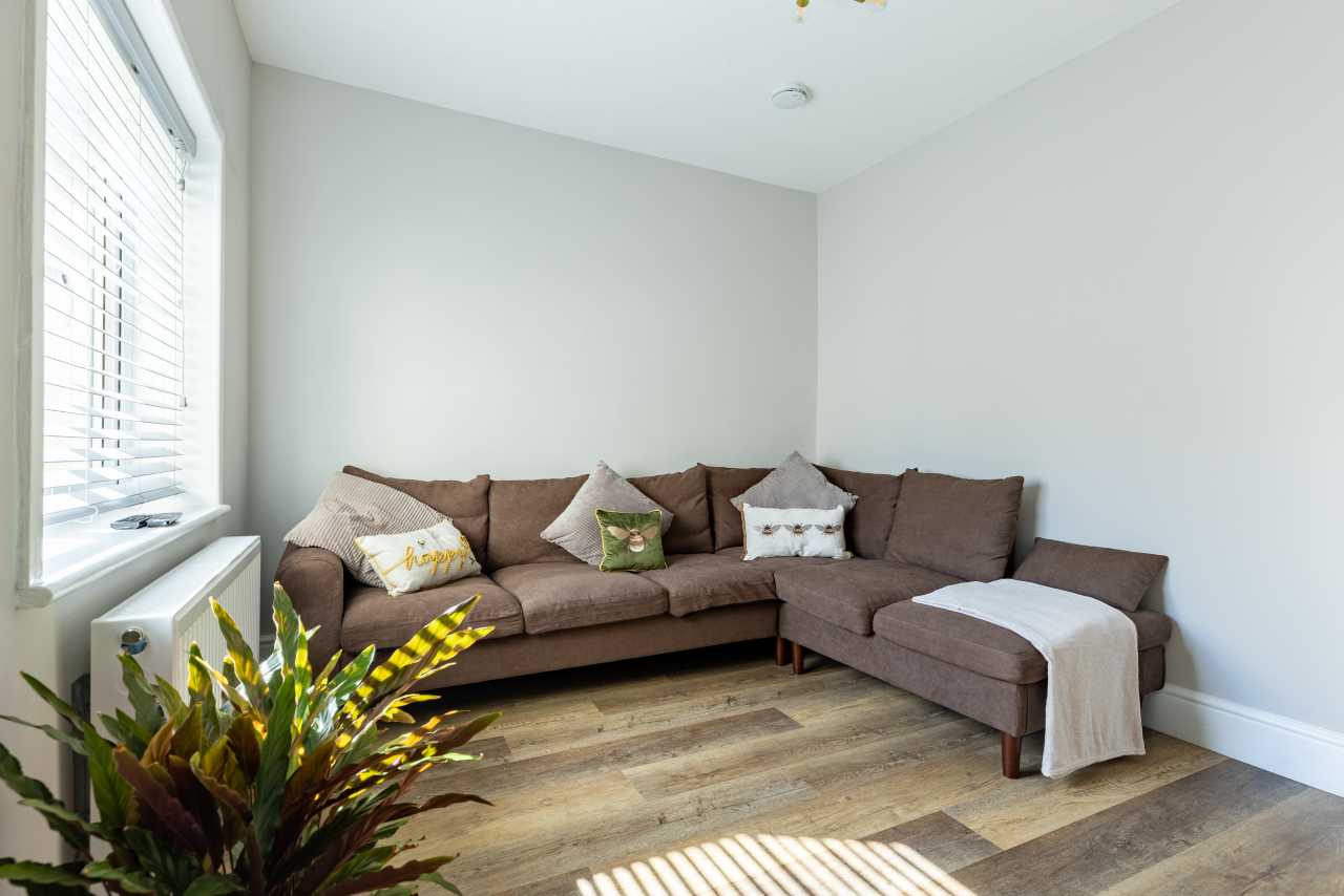 Bright sitting area with an L-shaped sofa, cushions, a throw, houseplant, window blinds and wooden flooring.