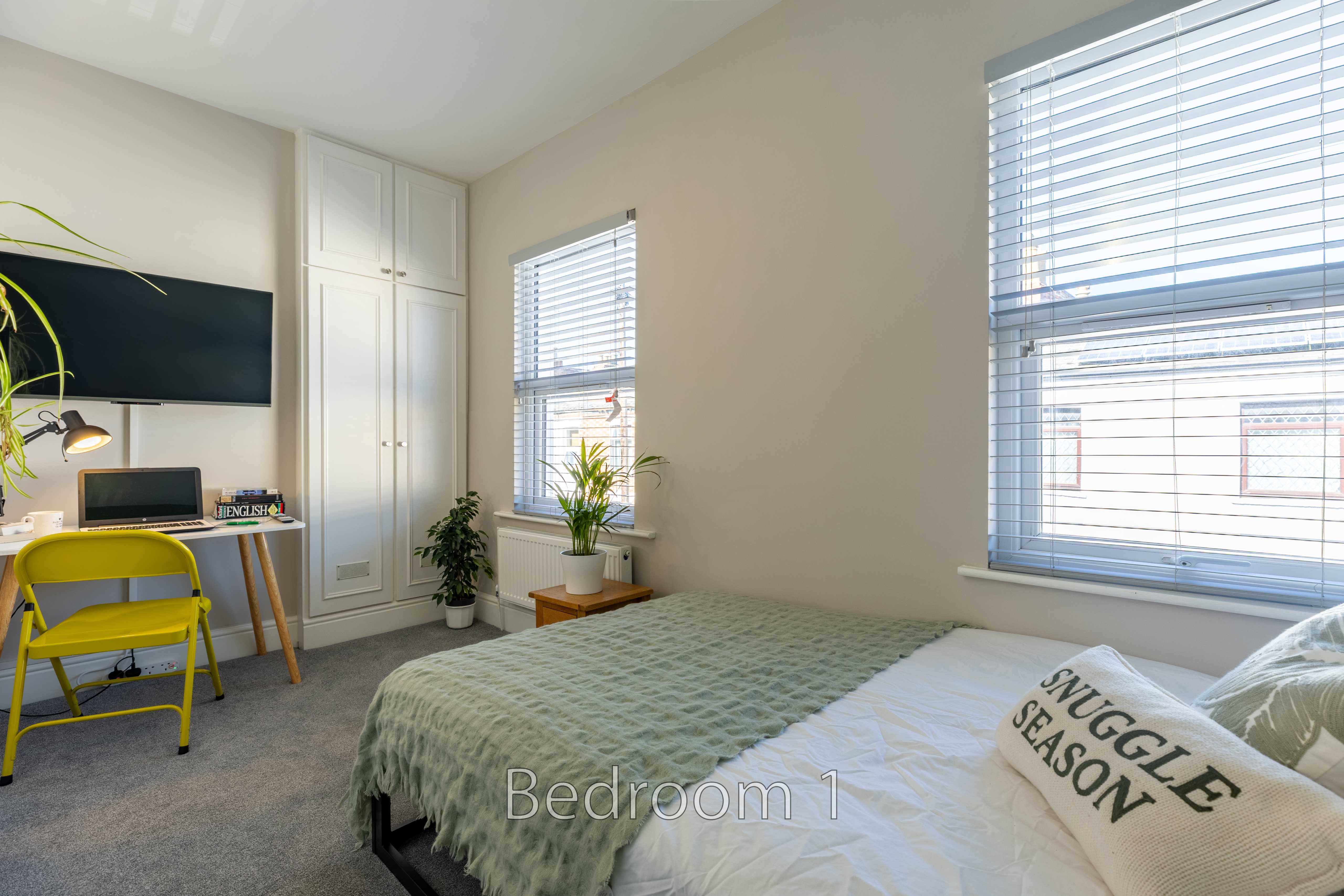 A bright bedroom with a double bed, two windows with blinds, built-in storage, a desk and a yellow chair.