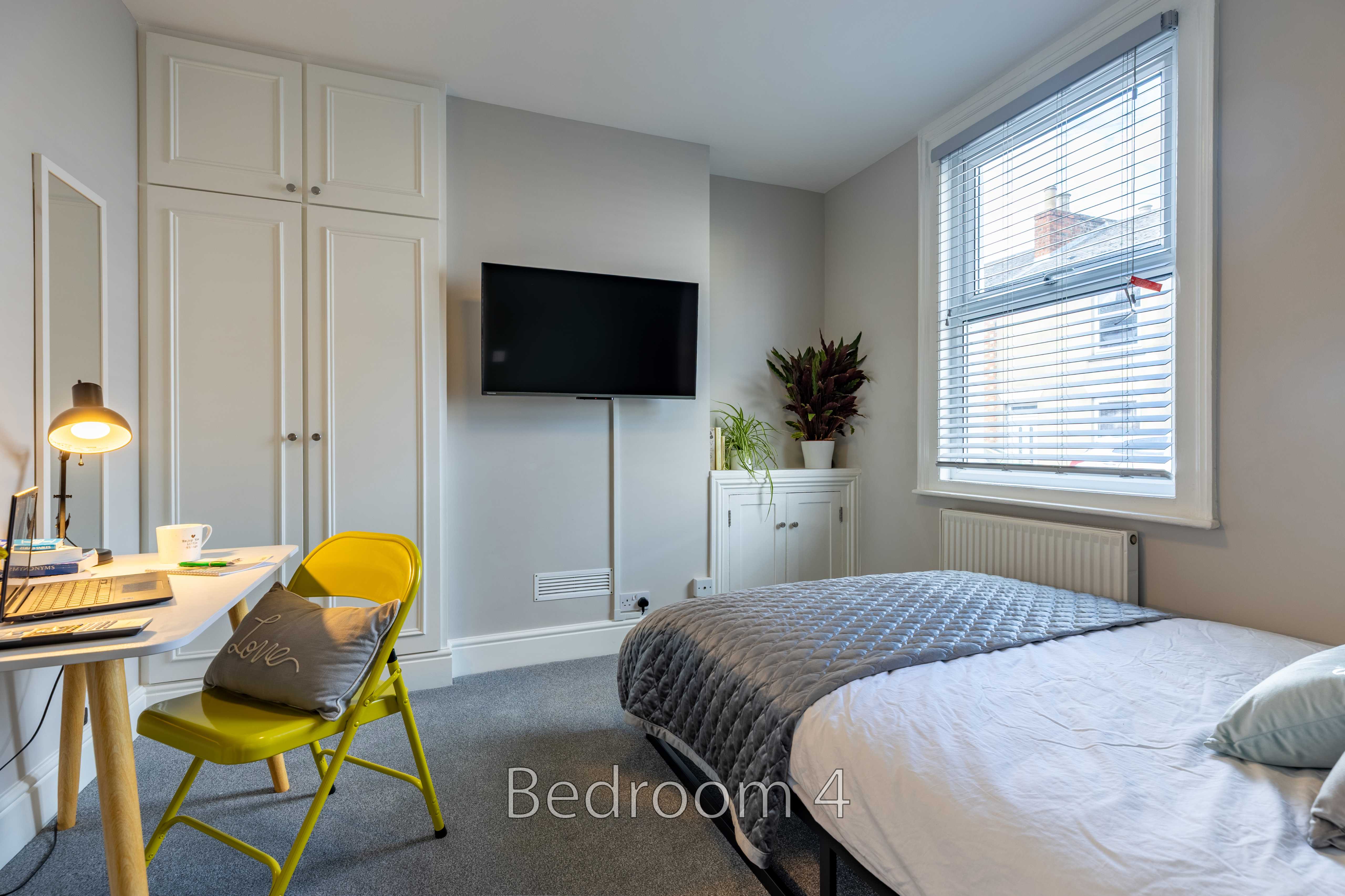 A furnished bedroom with a bed, wall-mounted television, desk, chair, storage cupboards, and a window with blinds.