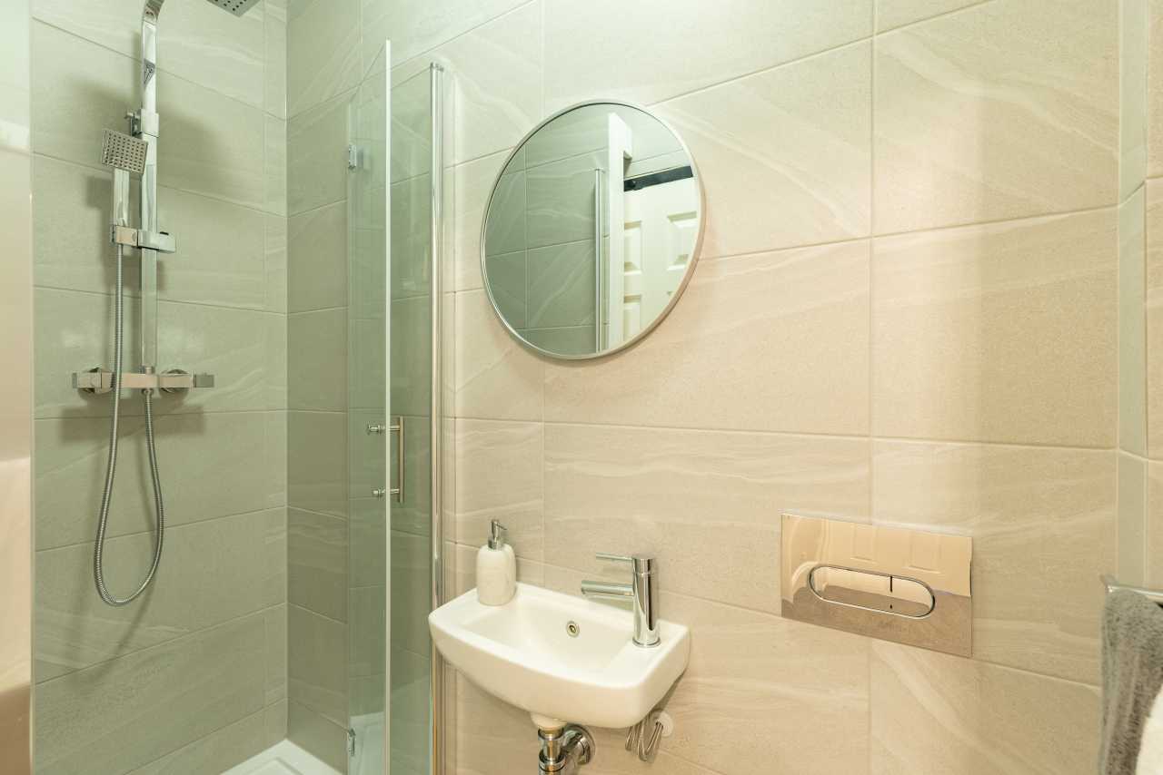 A tiled bathroom with a glass shower enclosure, wall-mounted sink, round mirror and toilet flush plate.