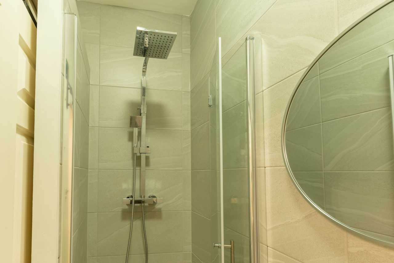 A tiled shower enclosure with a rainfall showerhead, handheld shower, glass door and large round wall mirror.