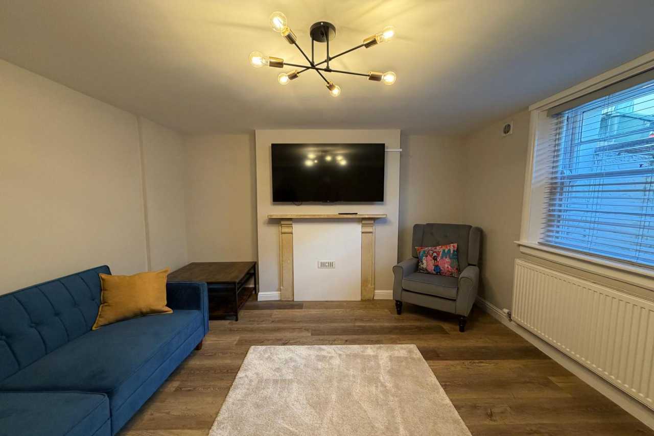 Furnished living room with a blue sofa, armchair, television above a fireplace, rug, window and radiator.