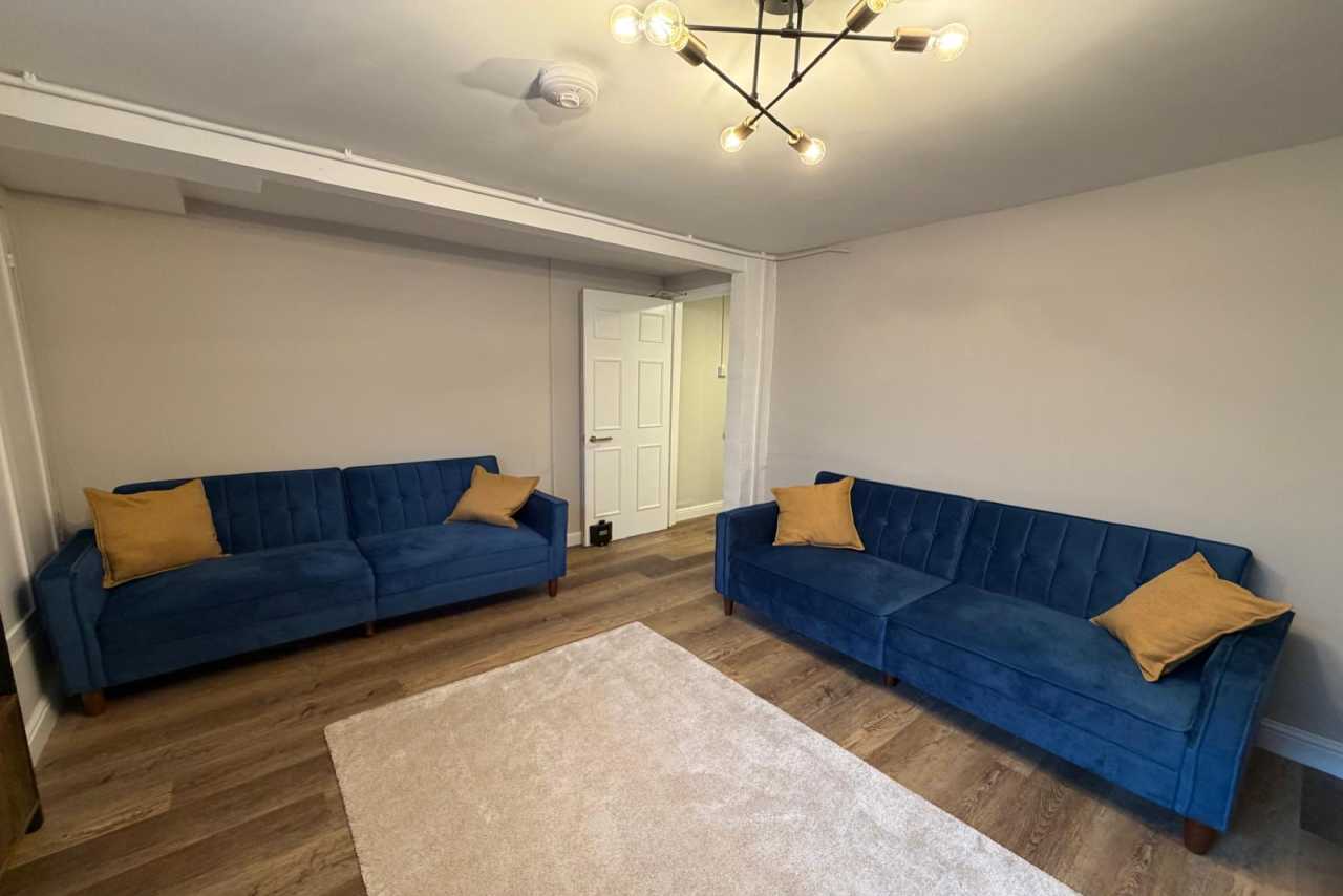 A furnished living room with two blue sofas, yellow cushions, a large rug, wood flooring and a modern ceiling light.