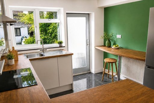 Bright modern kitchen with wood countertops, white cabinets, a sink beneath a window, a breakfast bar, stool and green accent wall.