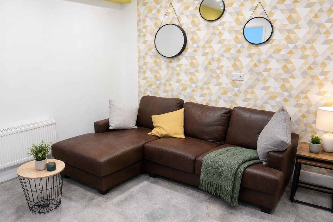 A furnished lounge with a large sectional sofa, patterned accent wall, round mirrors, side tables, plants and a radiator.