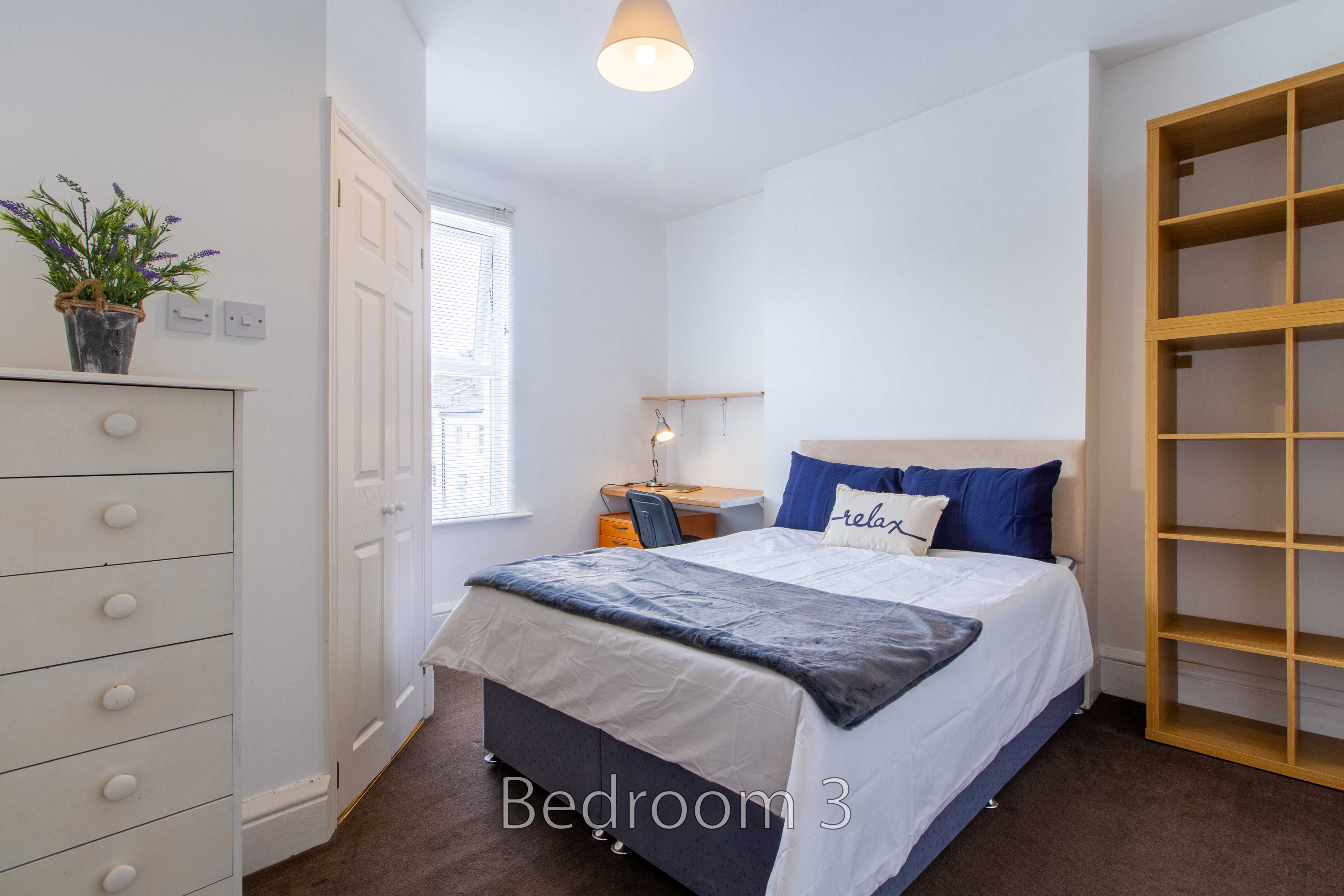 A furnished bedroom with a double bed, bedside table, chest of drawers, wardrobe shelving and a window.